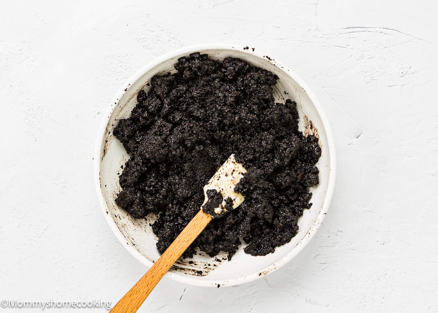 A white bowl filled with crushed Oreo cookies and cream mixture for a No Bake Oreo Cheesecake Pie, with a wooden spatula resting inside, on a white textured surface.