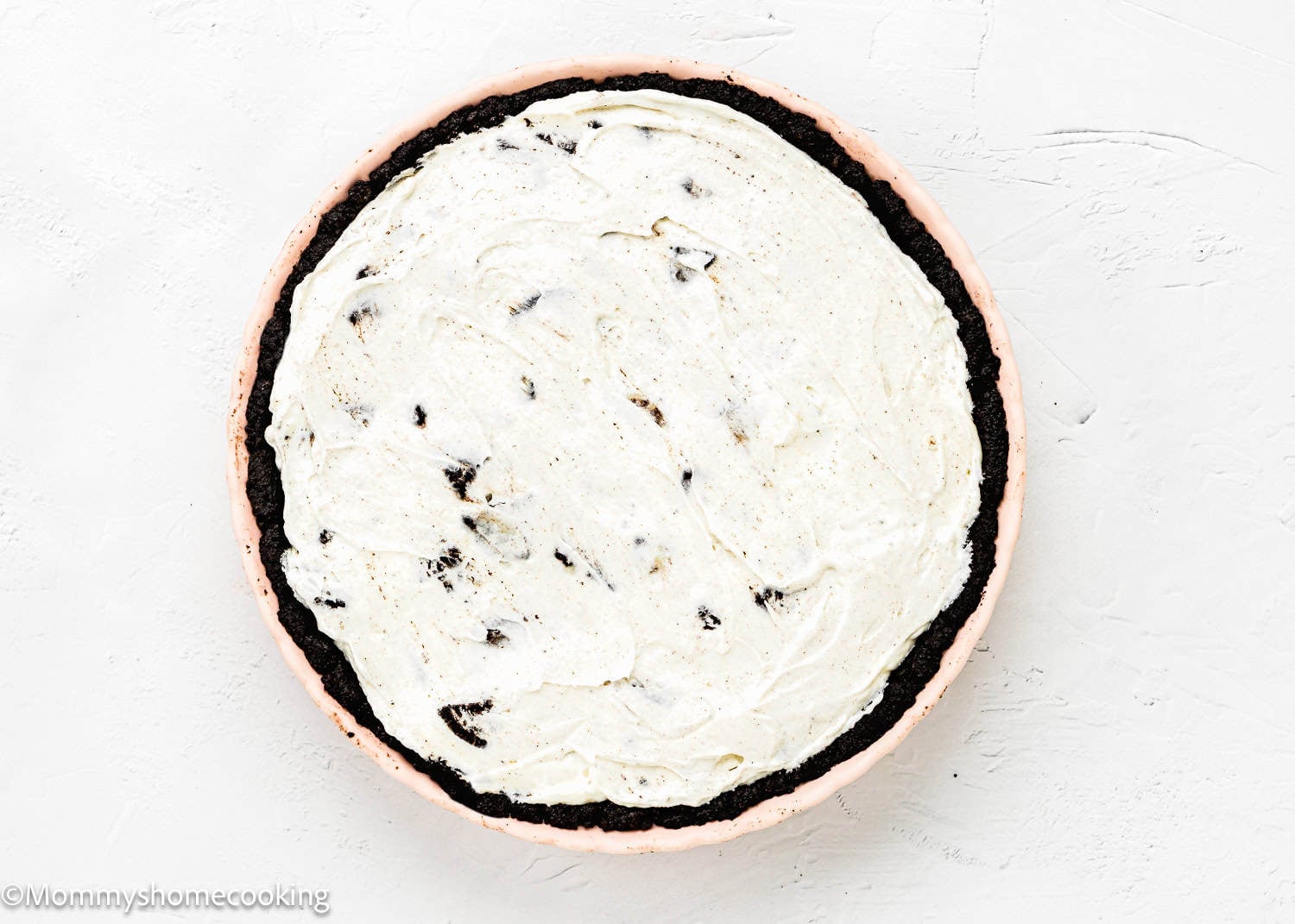 A No Bake Oreo Cheesecake Pie with a dark chocolate crust and creamy white filling is shown in a light pink pie dish on a white surface.