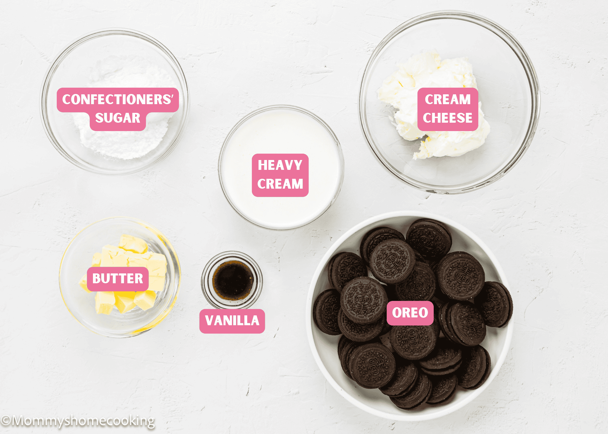 Five bowls on a white surface hold labeled ingredients for No Bake Oreo Cheesecake Pie: confectioners’ sugar, cream cheese, heavy cream, butter, vanilla extract, and a bowl of Oreo cookies.
