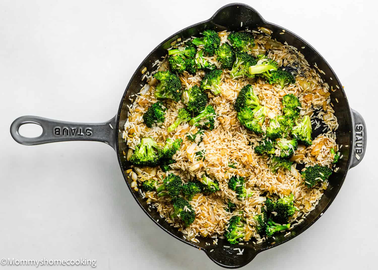 A black skillet contains uncooked rice, broccoli florets, and onion pieces on a white surface, ready to become a flavorful One Pot Chicken Broccoli Rice Casserole.