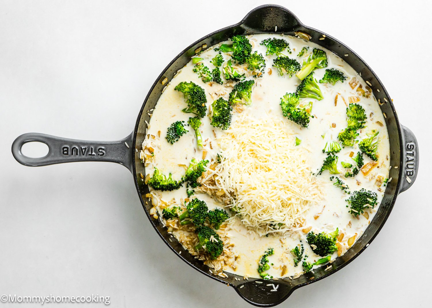 A cast iron skillet filled with broccoli, shredded cheese, creamy white sauce, and all the makings of a One Pot Chicken Broccoli Rice Casserole, ready to be cooked.