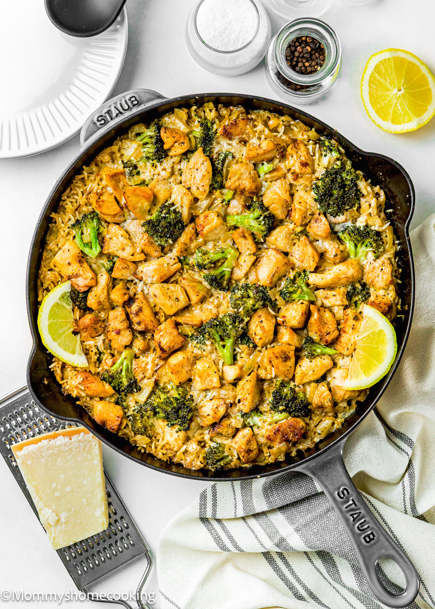 Skillet with One Pot Chicken Broccoli Rice Casserole, garnished with lemon slices; cheese grater, plate, and seasonings nearby on a white surface.