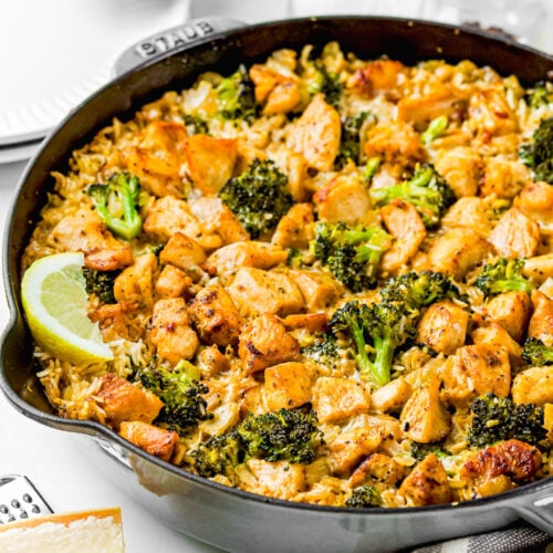 A skillet filled with One Pot Chicken Broccoli Rice Casserole—cooked rice, broccoli, and seasoned chicken pieces garnished with a lemon wedge—sits on a white surface next to kitchen utensils and plates.