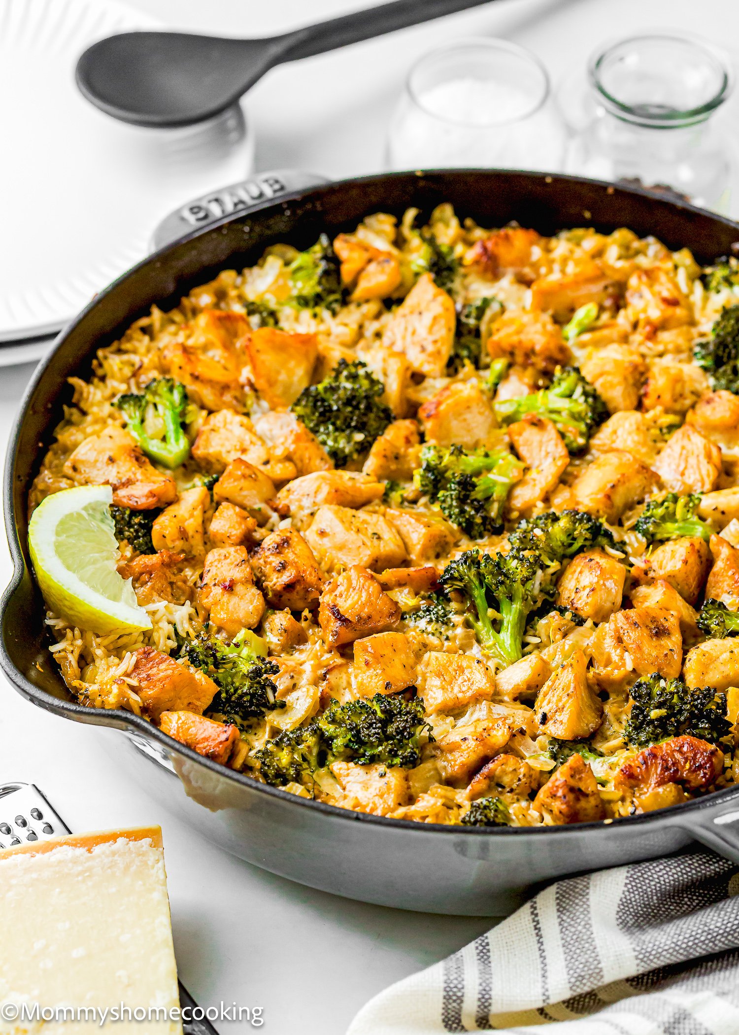 A skillet filled with One Pot Chicken Broccoli Rice Casserole—cooked rice, broccoli, and seasoned chicken pieces garnished with a lemon wedge—sits on a white surface next to kitchen utensils and plates.