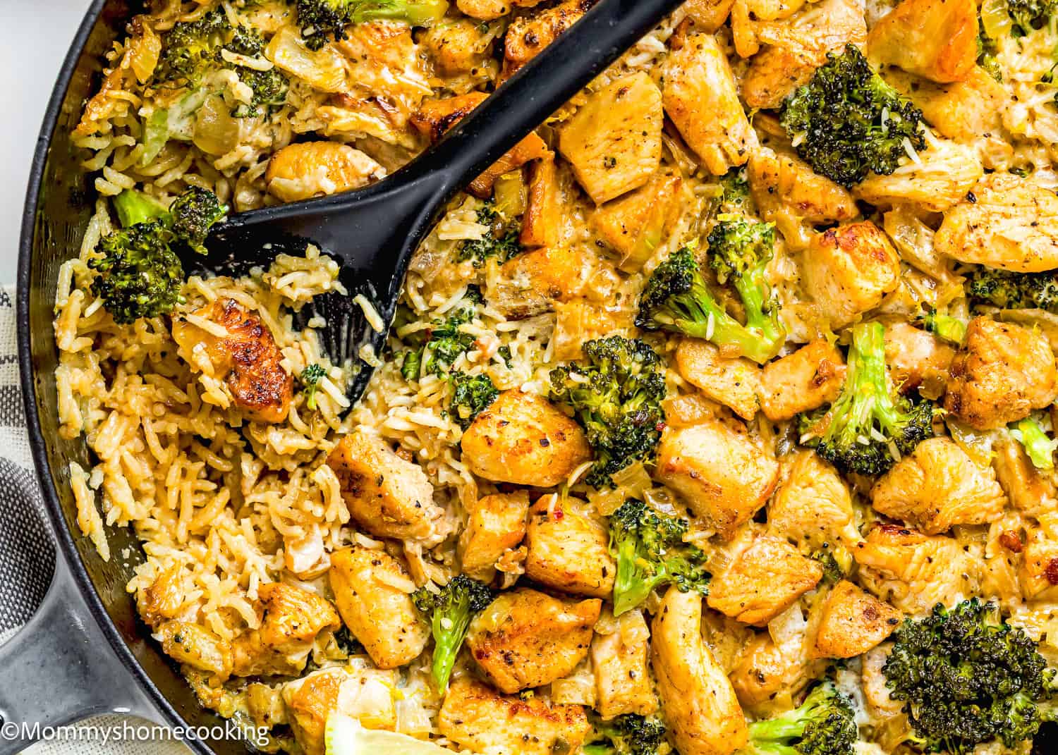 A skillet filled with One Pot Chicken Broccoli Rice Casserole, featuring cooked rice, broccoli florets, and seasoned chicken pieces, with a black serving spoon scooping some of the food.