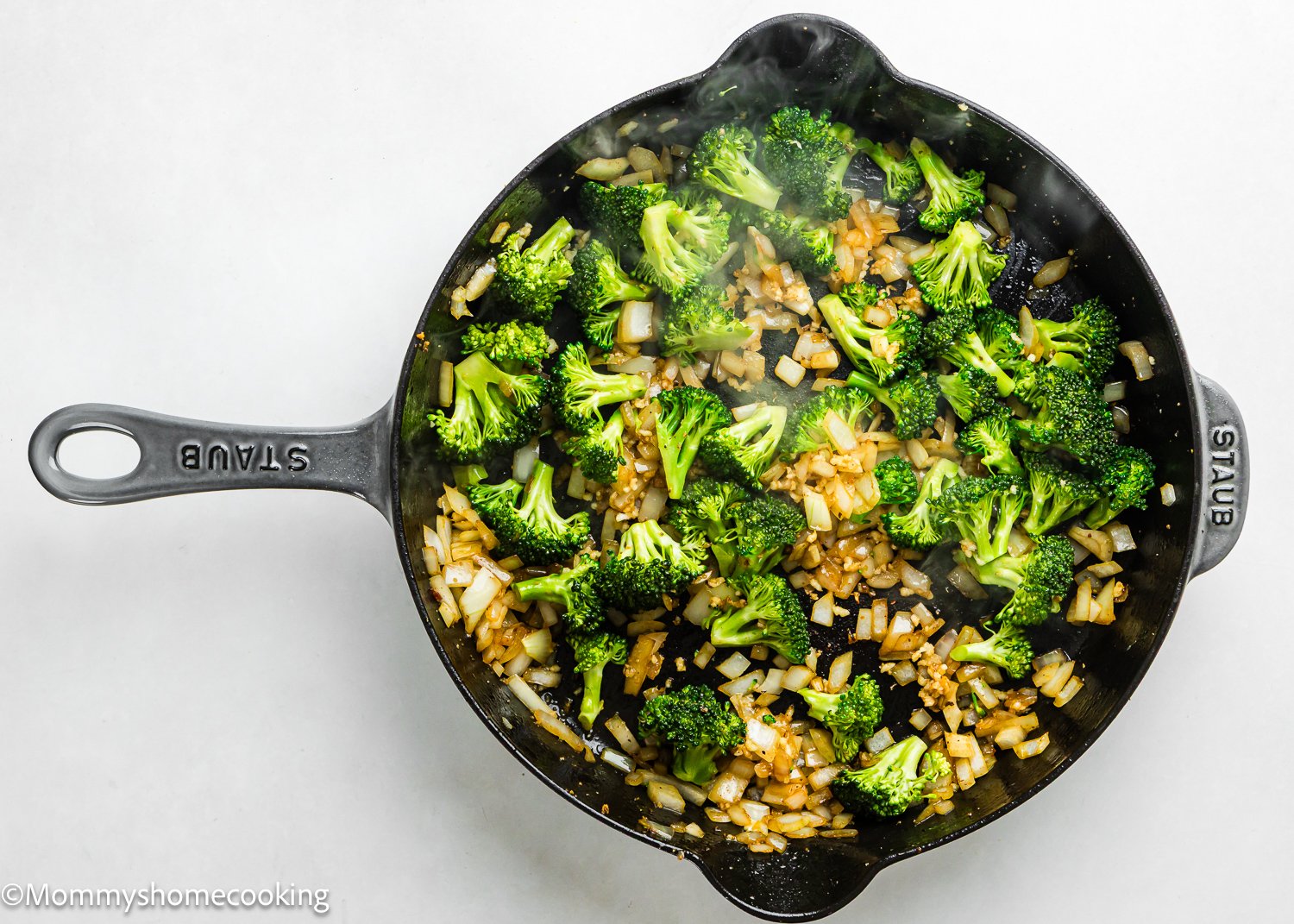 Overhead view of a cast iron skillet with cooked broccoli florets and diced onions on a white surface, perfect for adding to a One Pot Chicken Broccoli Rice Casserole.