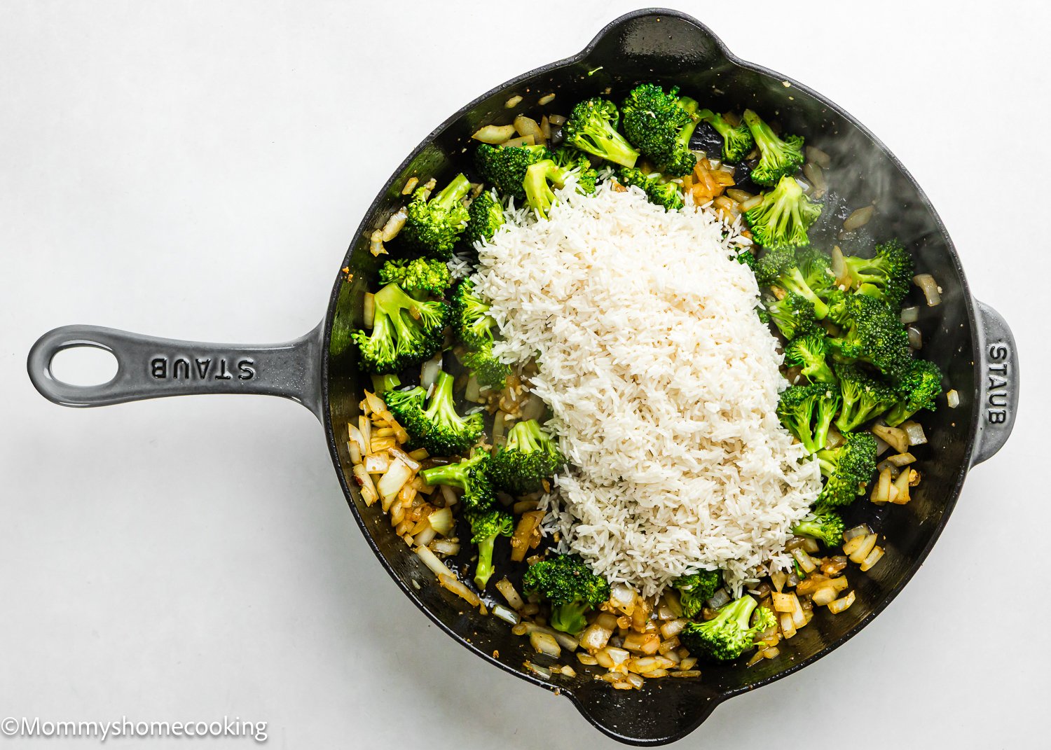 A cast iron skillet containing chopped onions, broccoli florets, and a mound of uncooked rice on a white surface—perfect for making One Pot Chicken Broccoli Rice Casserole.