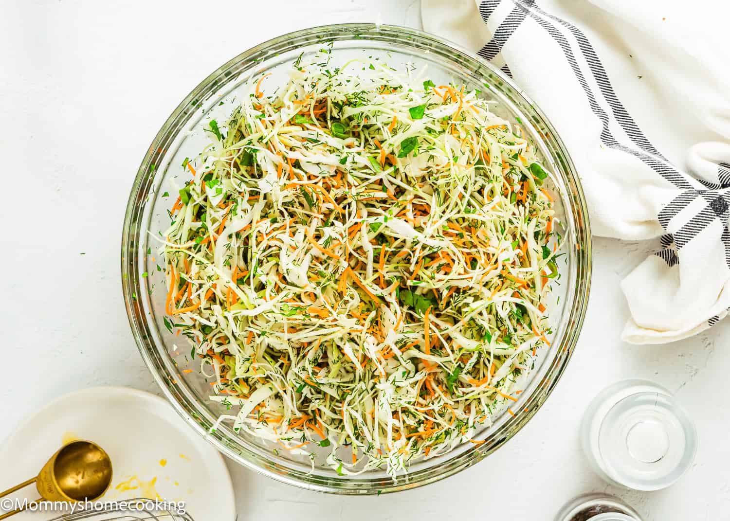 A clear glass bowl filled with Simple Cabbage Salad (No Mayo)—shredded cabbage, carrots, and herbs—sits on a white surface beside a striped towel and kitchen utensils.