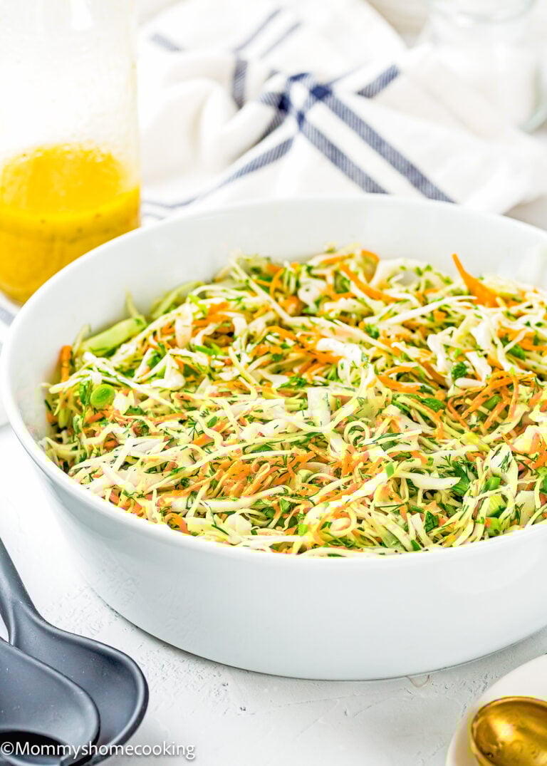 A white bowl filled with Simple Cabbage Salad (No Mayo), made of shredded cabbage and carrots, sits on a white surface with a jar of yellow dressing and utensils nearby.