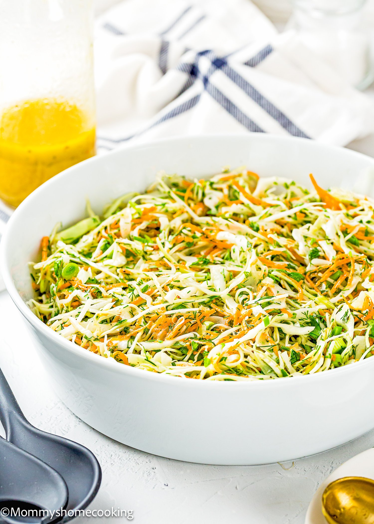 A white bowl filled with Simple Cabbage Salad (No Mayo), made of shredded cabbage and carrots, sits on a white surface with a jar of yellow dressing and utensils nearby.