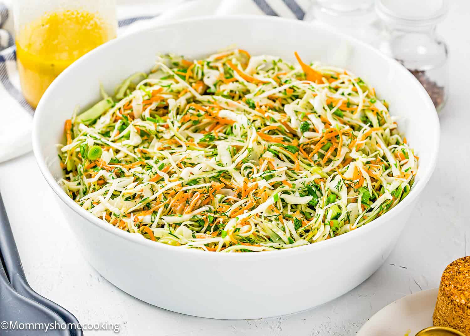 A white bowl filled with shredded cabbage, carrots, and green onions creates a fresh Simple Cabbage Salad (No Mayo). A jar of dressing and seasonings is in the background.