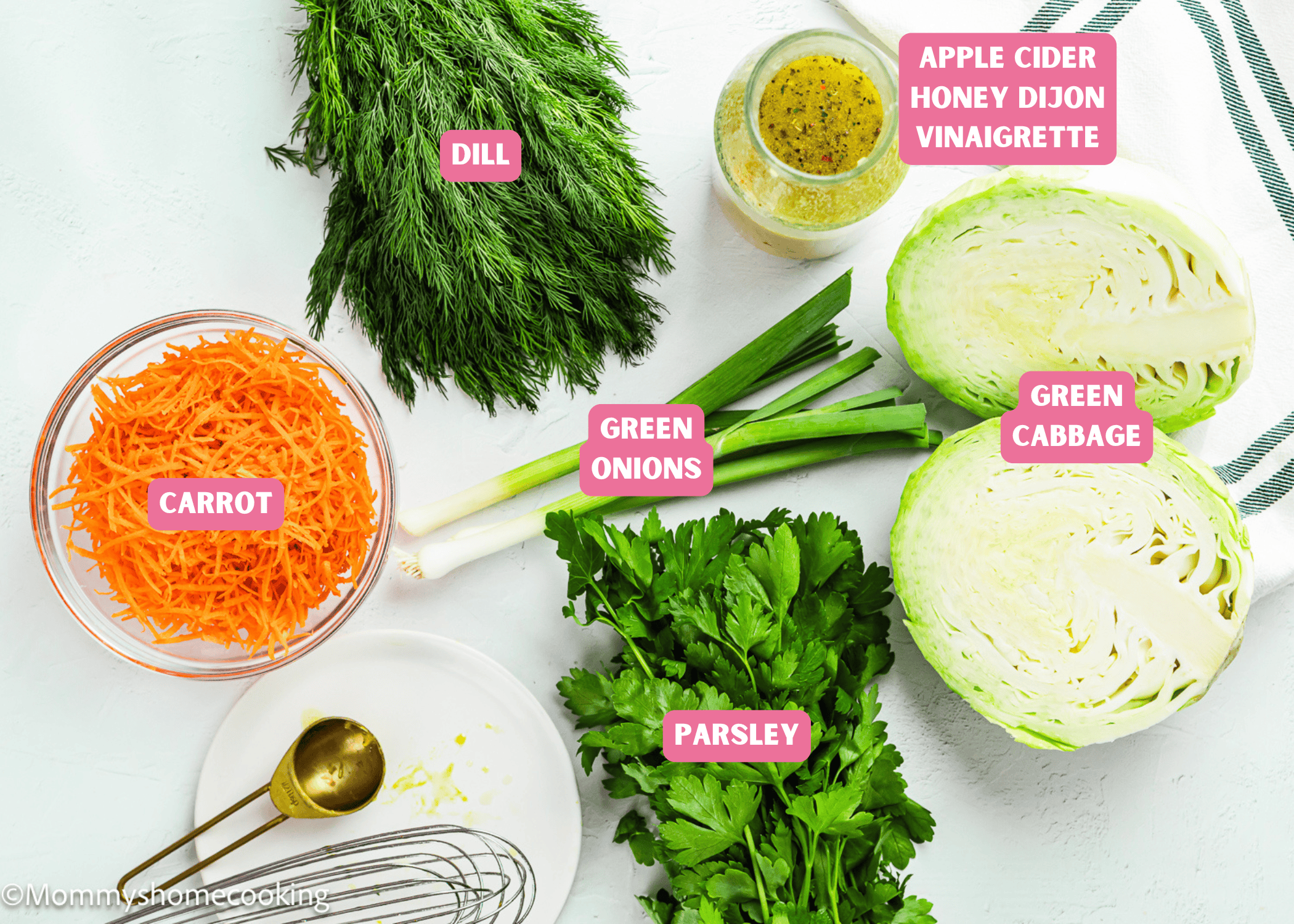 Assorted ingredients on a white surface, including shredded carrot, dill, green onions, parsley, green cabbage, and a jar of apple cider honey dijon vinaigrette for a Simple Cabbage Salad (No Mayo).