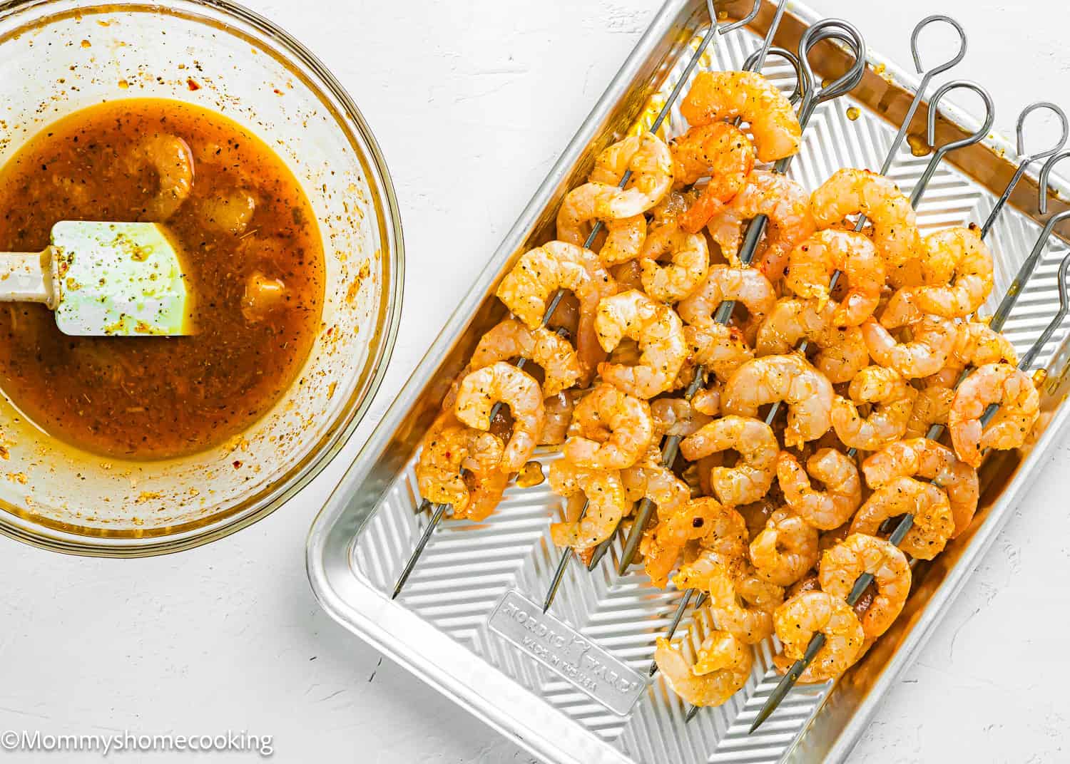 Raw shrimp coated in seasoning are arranged on metal skewers in a tray, with a bowl of Simple Shrimp Marinade (Perfect for Grilling!) and a spatula beside them on a white background.