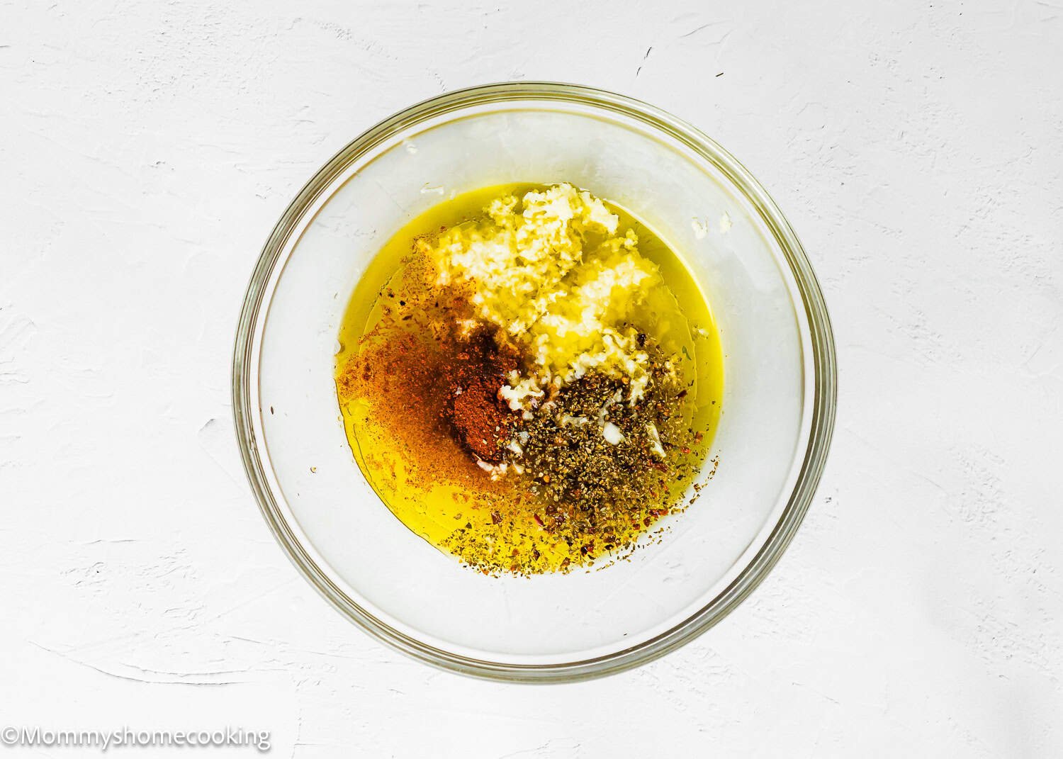 A glass bowl containing olive oil, minced garlic, black pepper, paprika, and dried herbs makes a Simple Shrimp Marinade (Perfect for Grilling!) on a white surface.