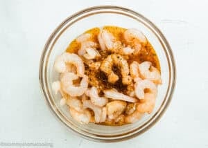 A glass bowl filled with raw shrimp marinating in a Simple Shrimp Marinade (Perfect for Grilling!) sits on a white surface.