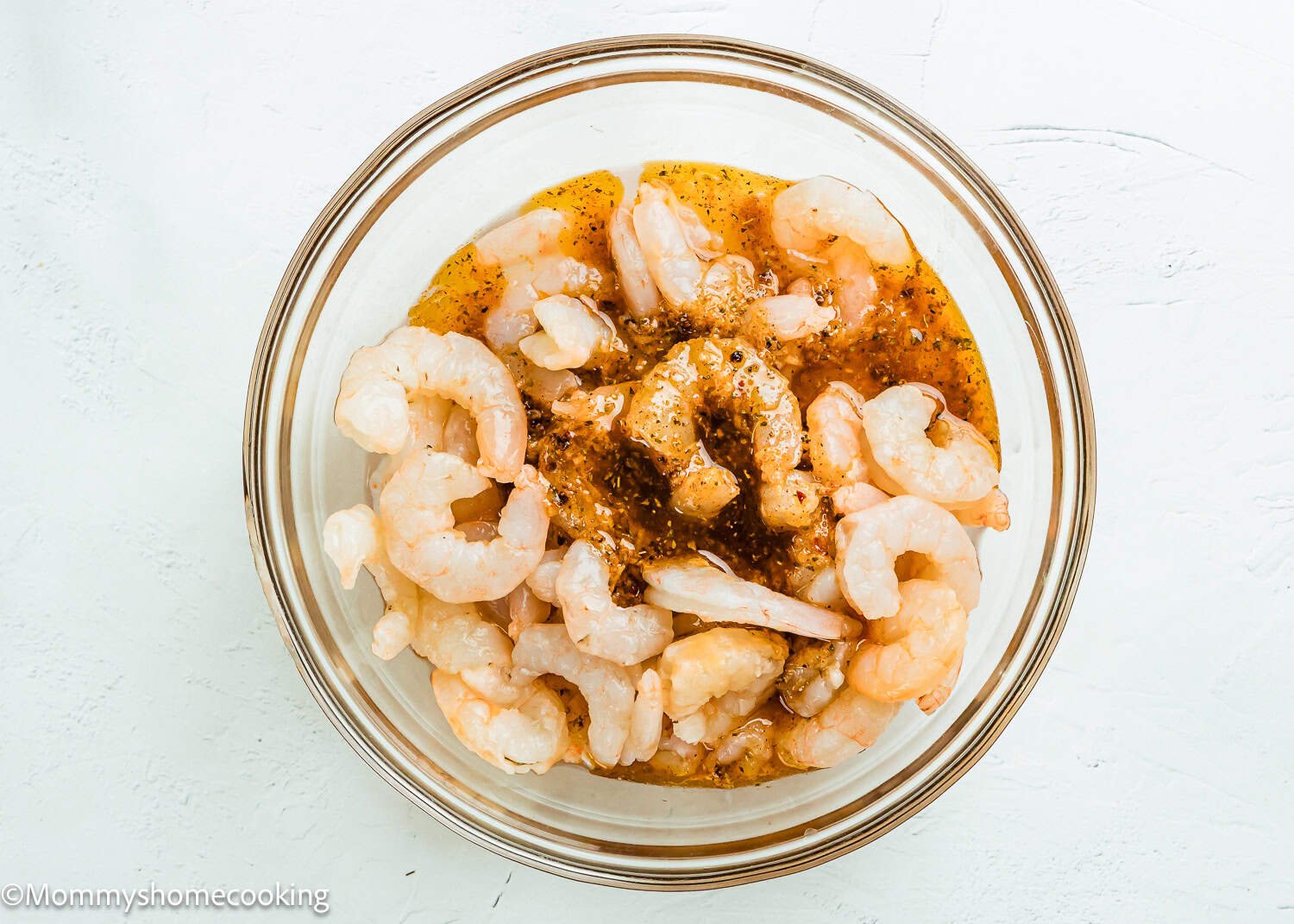 A glass bowl filled with raw shrimp marinating in a Simple Shrimp Marinade (Perfect for Grilling!) sits on a white surface.