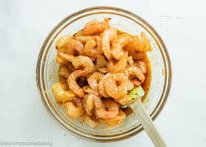 A glass bowl filled with raw shrimp tossed in a Simple Shrimp Marinade (Perfect for Grilling!) is being mixed with a green spatula on a white surface.