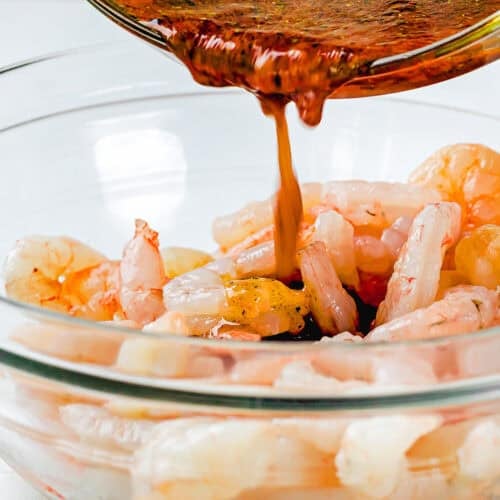 A bowl of raw shrimp is being marinated as a reddish-brown Simple Shrimp Marinade (Perfect for Grilling!) is poured over them from a glass bowl.