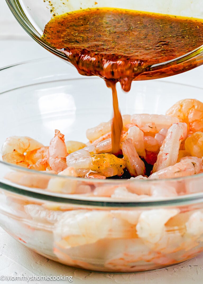 A bowl of raw shrimp is being marinated as a reddish-brown Simple Shrimp Marinade (Perfect for Grilling!) is poured over them from a glass bowl.