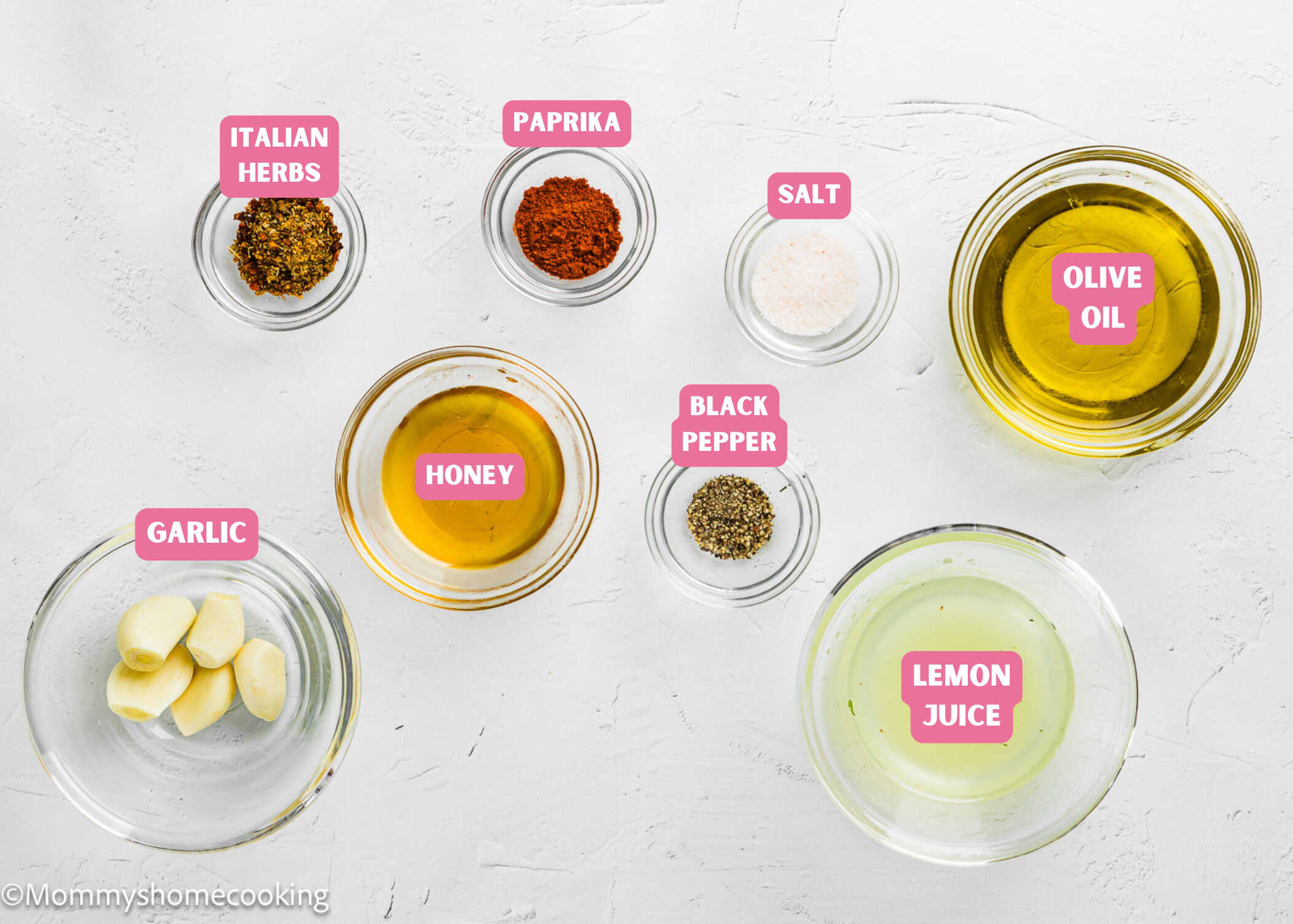 Top-down view of small bowls containing olive oil, lemon juice, honey, garlic cloves, Italian herbs, paprika, black pepper, and salt—ingredients for a Simple Shrimp Marinade (Perfect for Grilling!)—all labeled on a white surface.