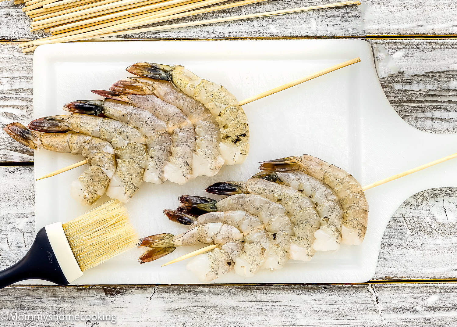 Raw shrimp skewered on wooden sticks are arranged on a white cutting board, with a basting brush beside them and more skewers in the background, ready for Super Easy Grilled Barbecue Shrimp.