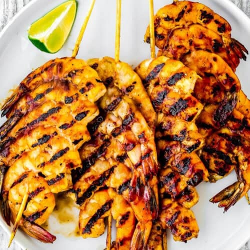 Super Easy Grilled Barbecue Shrimp skewers arranged on a white plate with grill marks, served with a wedge of lime.