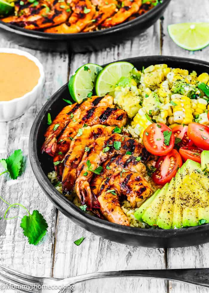 A bowl with Super Easy Grilled Barbecue Shrimp, sliced avocado, cherry tomatoes, corn, rice, lime wedges, and chopped herbs, served with a small dish of sauce on the side.