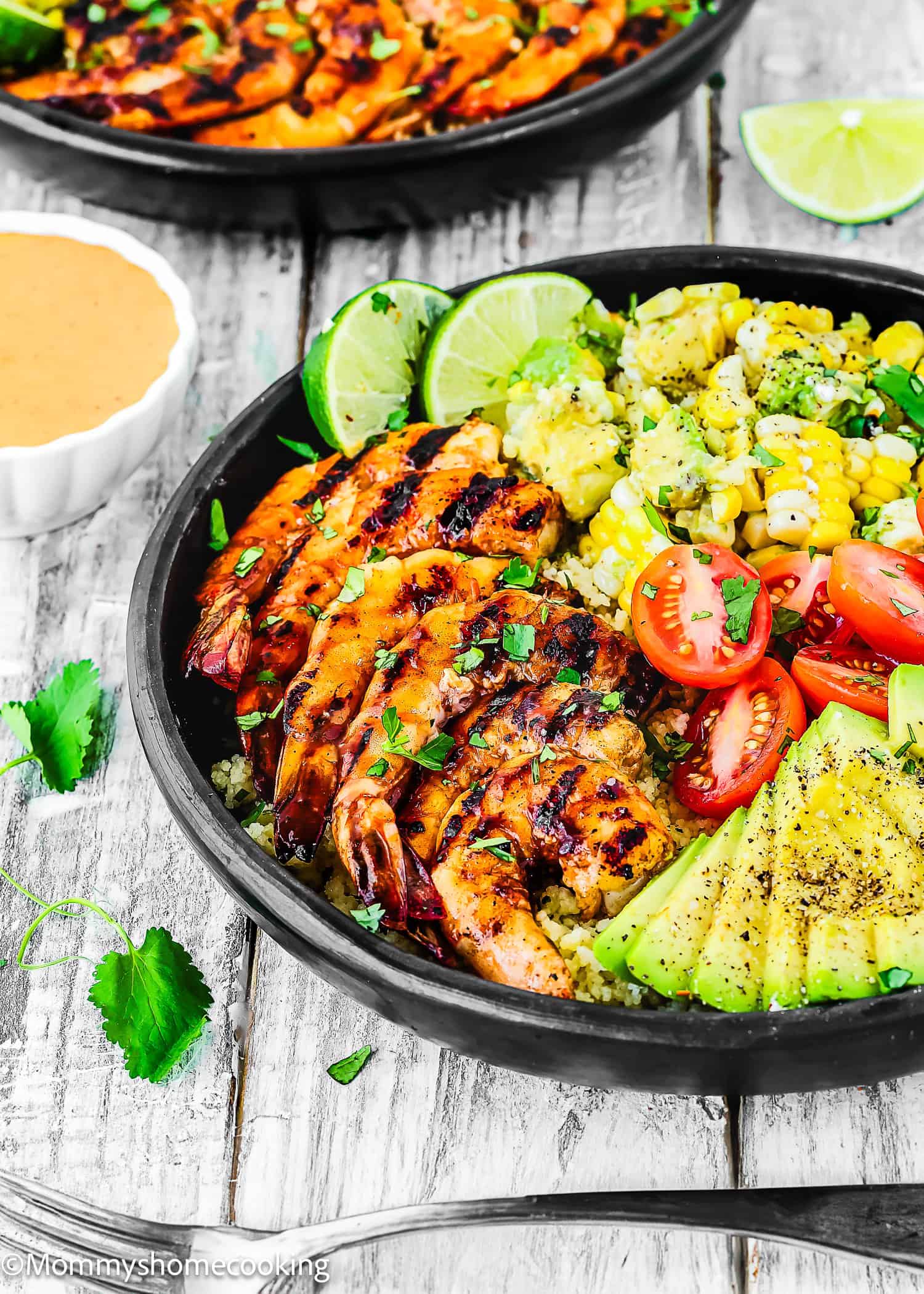 A bowl with Super Easy Grilled Barbecue Shrimp, sliced avocado, cherry tomatoes, corn, rice, lime wedges, and chopped herbs, served with a small dish of sauce on the side.