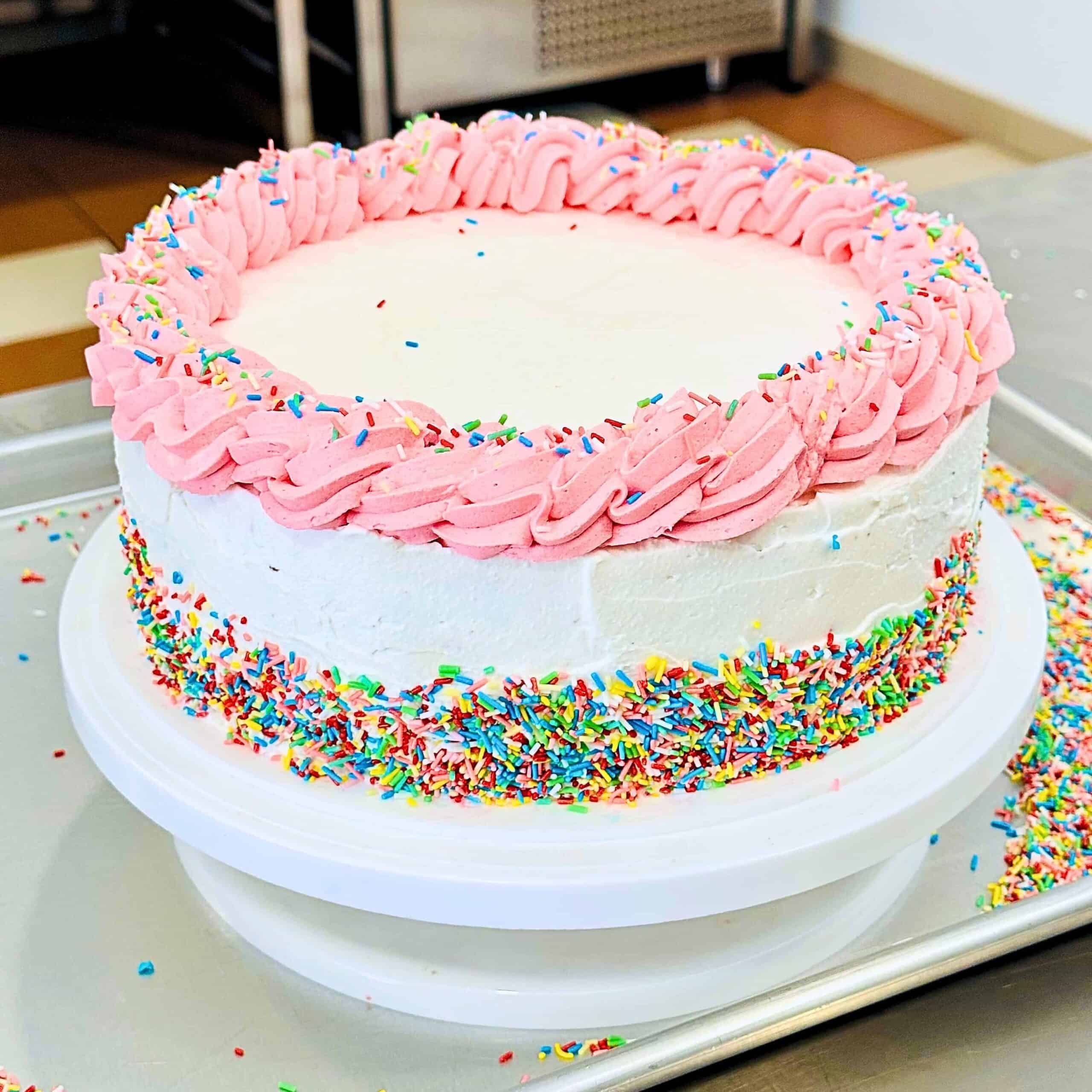 An eggless ice cream layer cake with white frosting, a pink piped border, and colorful sprinkles around the base sits on a white cake stand in a kitchen.