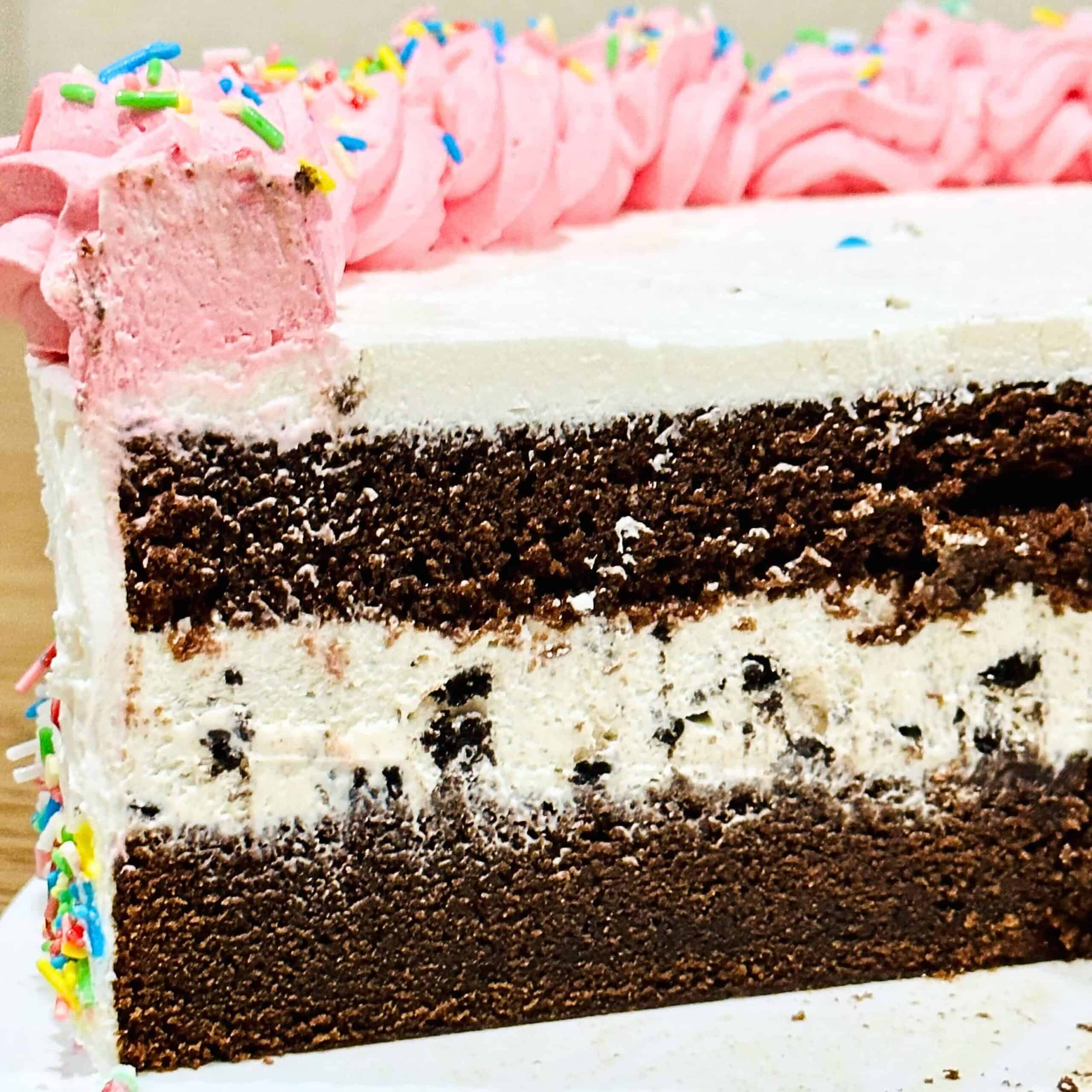 A close-up of a sliced eggless ice cream layer cake with chocolate and vanilla, pink frosting, colorful sprinkles, and a portion already cut out.