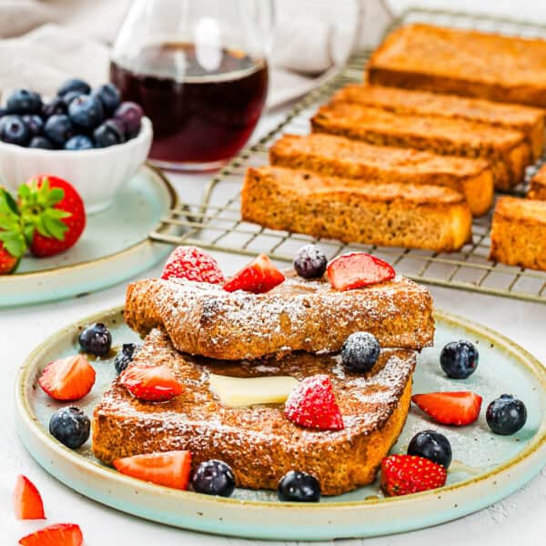 Two slices of Air Fryer Eggless French Toasts topped with powdered sugar, butter, strawberries, and blueberries on a plate, served with extra berries, syrup, and additional toast in the background.