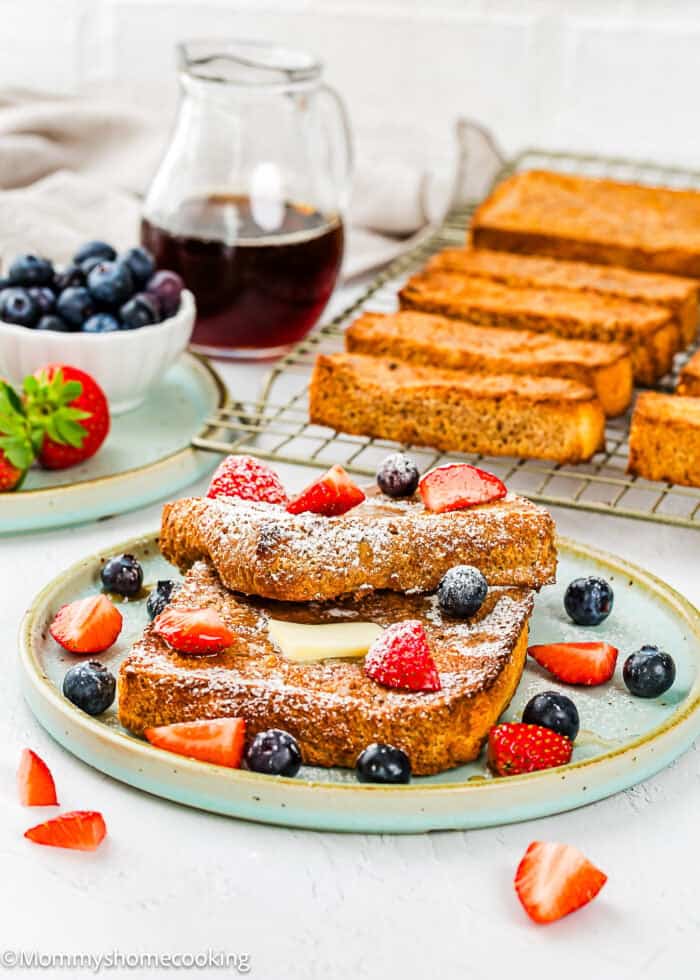 Two slices of Air Fryer Eggless French Toasts topped with powdered sugar, butter, strawberries, and blueberries on a plate, served with extra berries, syrup, and additional toast in the background.