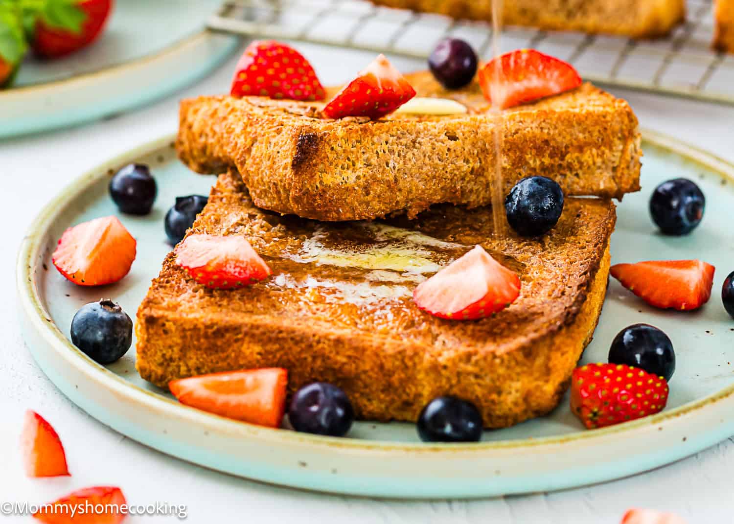 Two slices of Air Fryer Eggless French Toasts on a plate, topped with syrup, butter, strawberries, and blueberries. Additional fruit and toast are visible in the background.