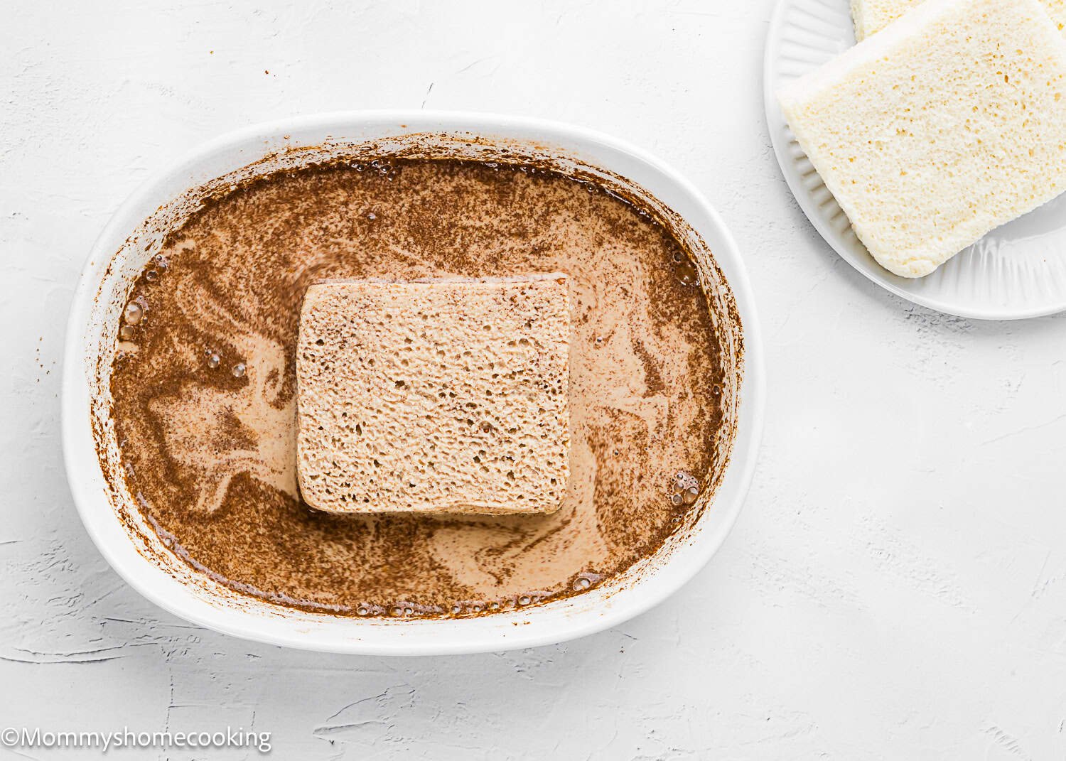 A slice of plain white bread soaking in a mixture of milk and cinnamon in a white oval dish, ready to be made into Air Fryer Eggless French Toasts, with more bread slices on a plate nearby.