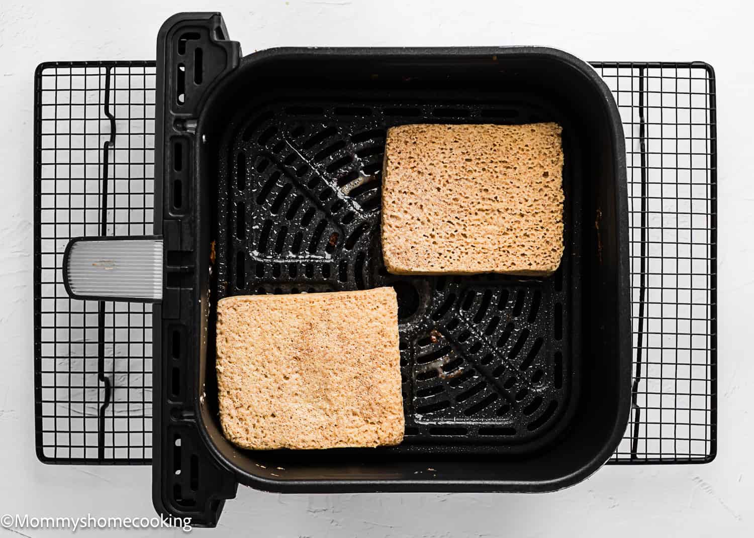 Two slices of bread are placed inside an air fryer basket, preparing to become delicious Air Fryer Eggless French Toasts on a cooling rack.