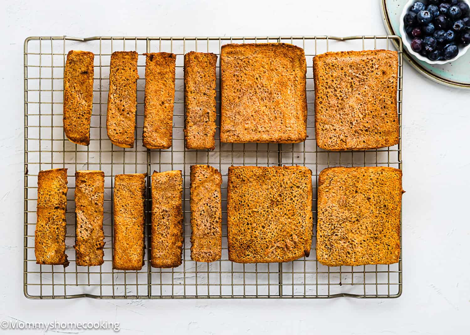 Slices and loaves of golden brown bread cooling on a wire rack, with a plate of blueberries in the upper right corner, perfect for making Air Fryer Eggless French Toasts.