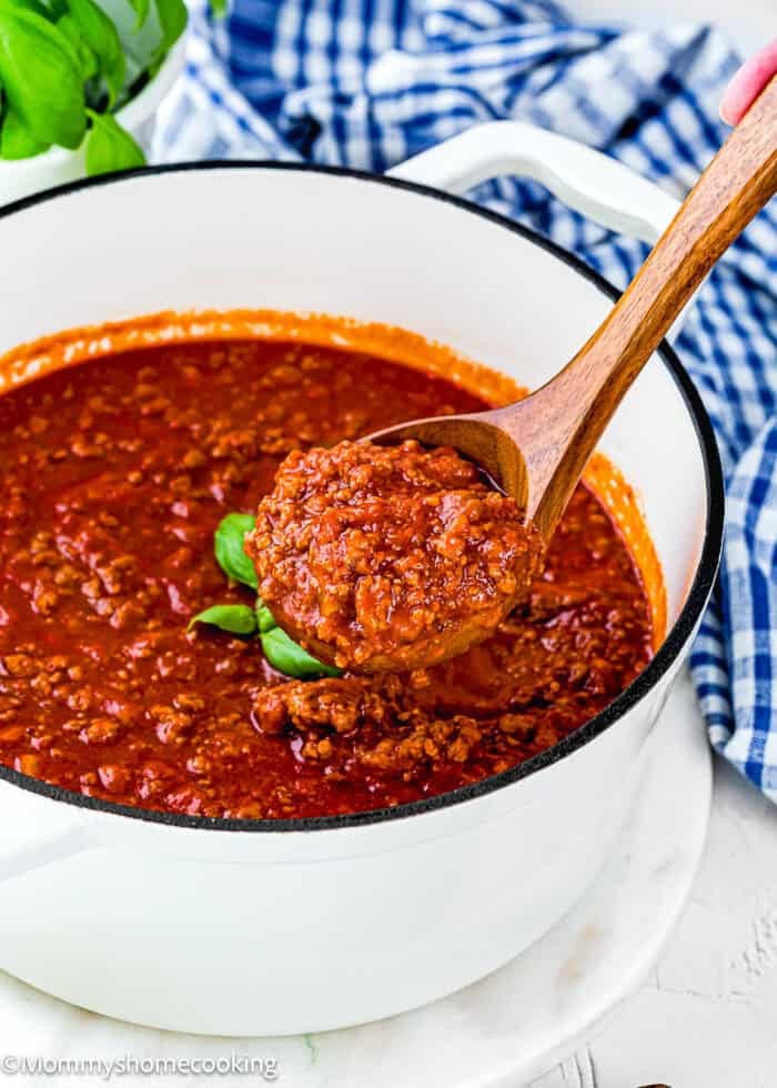 A wooden spoon lifts a portion of Classic Italian Bolognese Sauce from a white pot filled with chunky red tomato sauce, set on a marble surface with a blue checkered towel nearby.