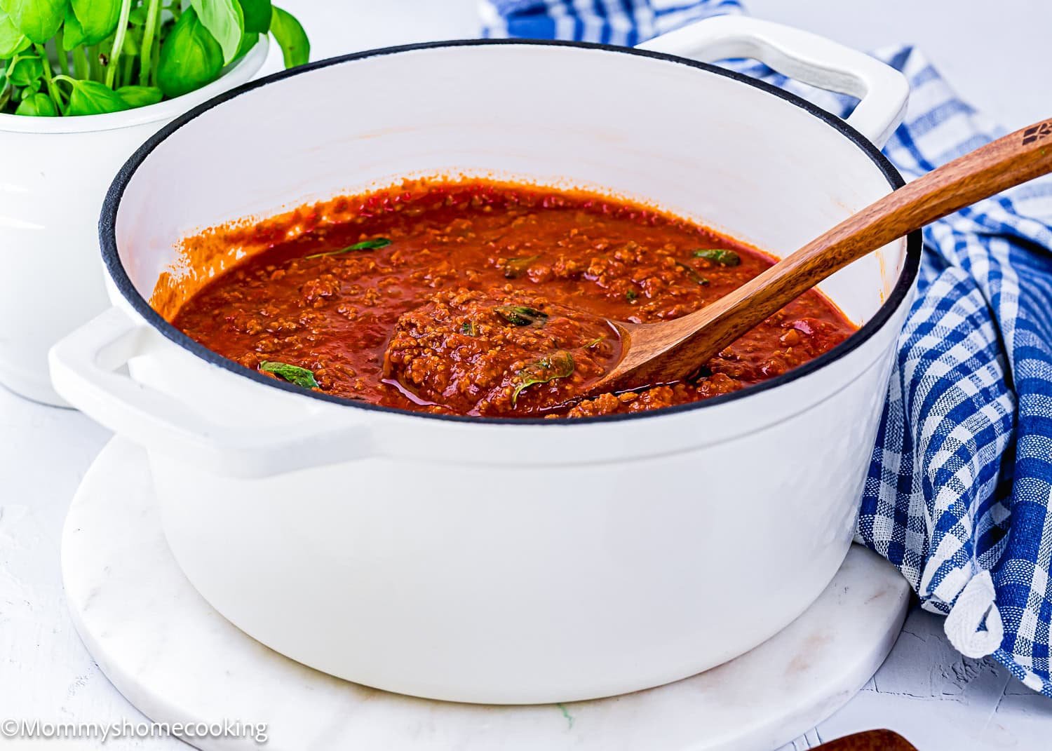 A white pot filled with Classic Italian Bolognese Sauce sits on a white surface, with a wooden spoon and a blue checkered cloth nearby.