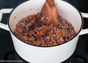 Ground beef, diced onions, and carrots are being cooked in a white pot with a wooden spoon to start a Classic Italian Bolognese Sauce.