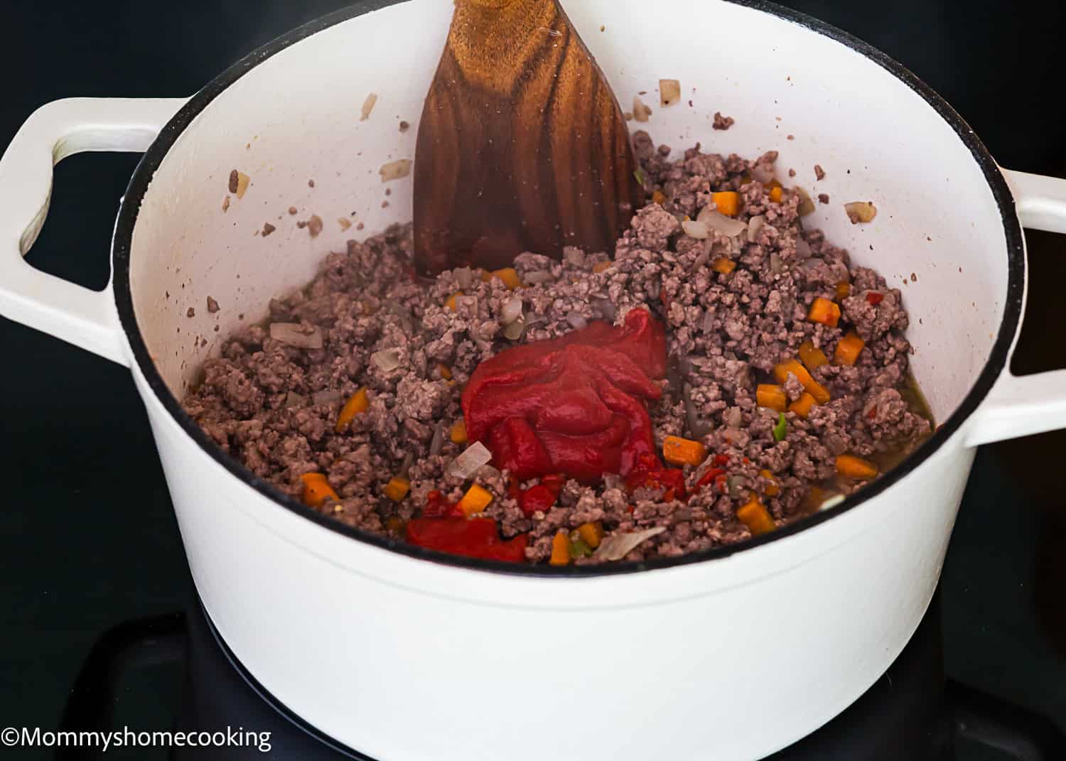 Ground beef, chopped onions, and carrots are being cooked in a white pot with tomato paste and stirred with a wooden spoon to create a Classic Italian Bolognese Sauce.