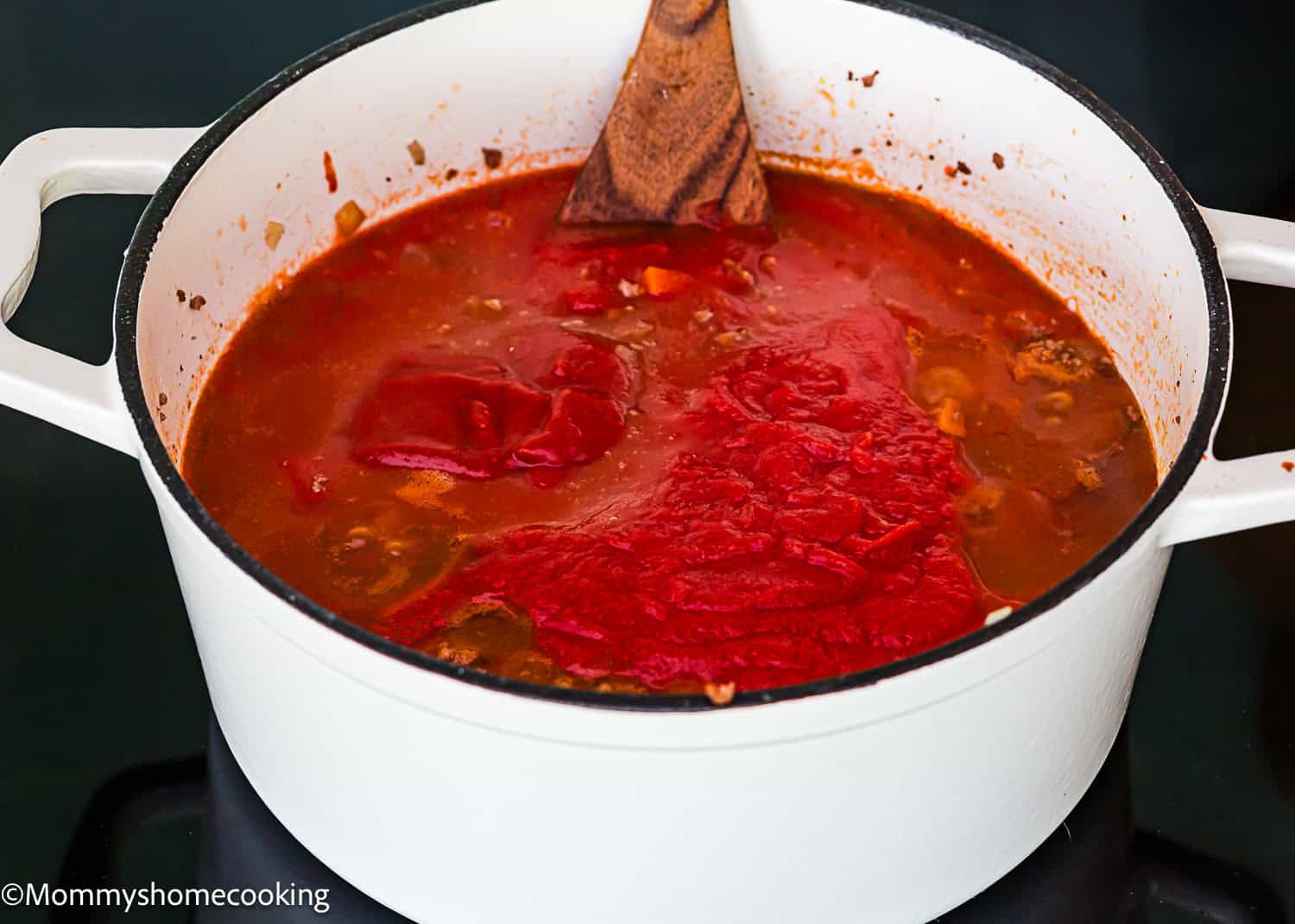 A white pot on a stove holds simmering classic Italian Bolognese sauce with chopped vegetables and a wooden spoon resting inside.