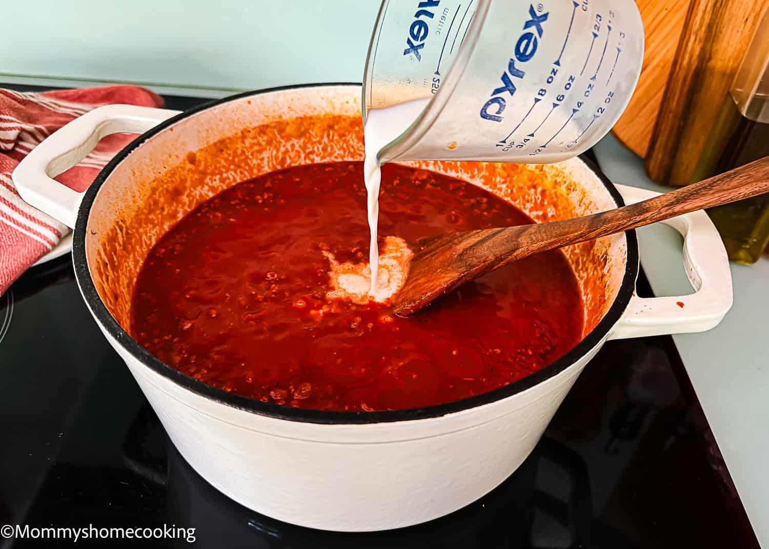 A hand pours a white liquid from a measuring cup into a pot of red Classic Italian Bolognese Sauce on a stovetop, with a wooden spoon resting inside the pot.