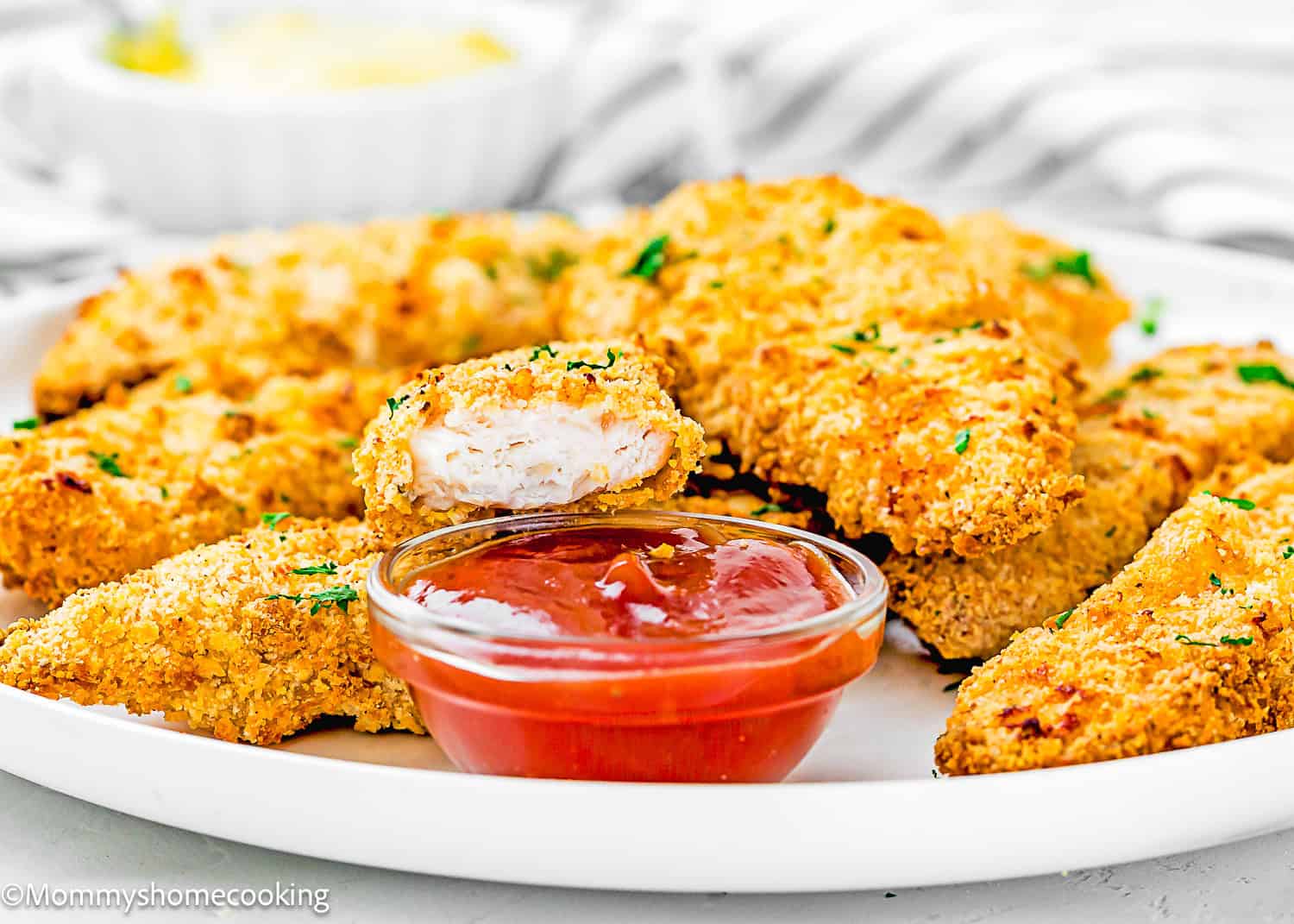 A plate of Easy Air Fryer Chicken Tenders (No Eggs - No Dairy) garnished with herbs, served with a small cup of red dipping sauce; one tender is sliced to show the juicy inside.
