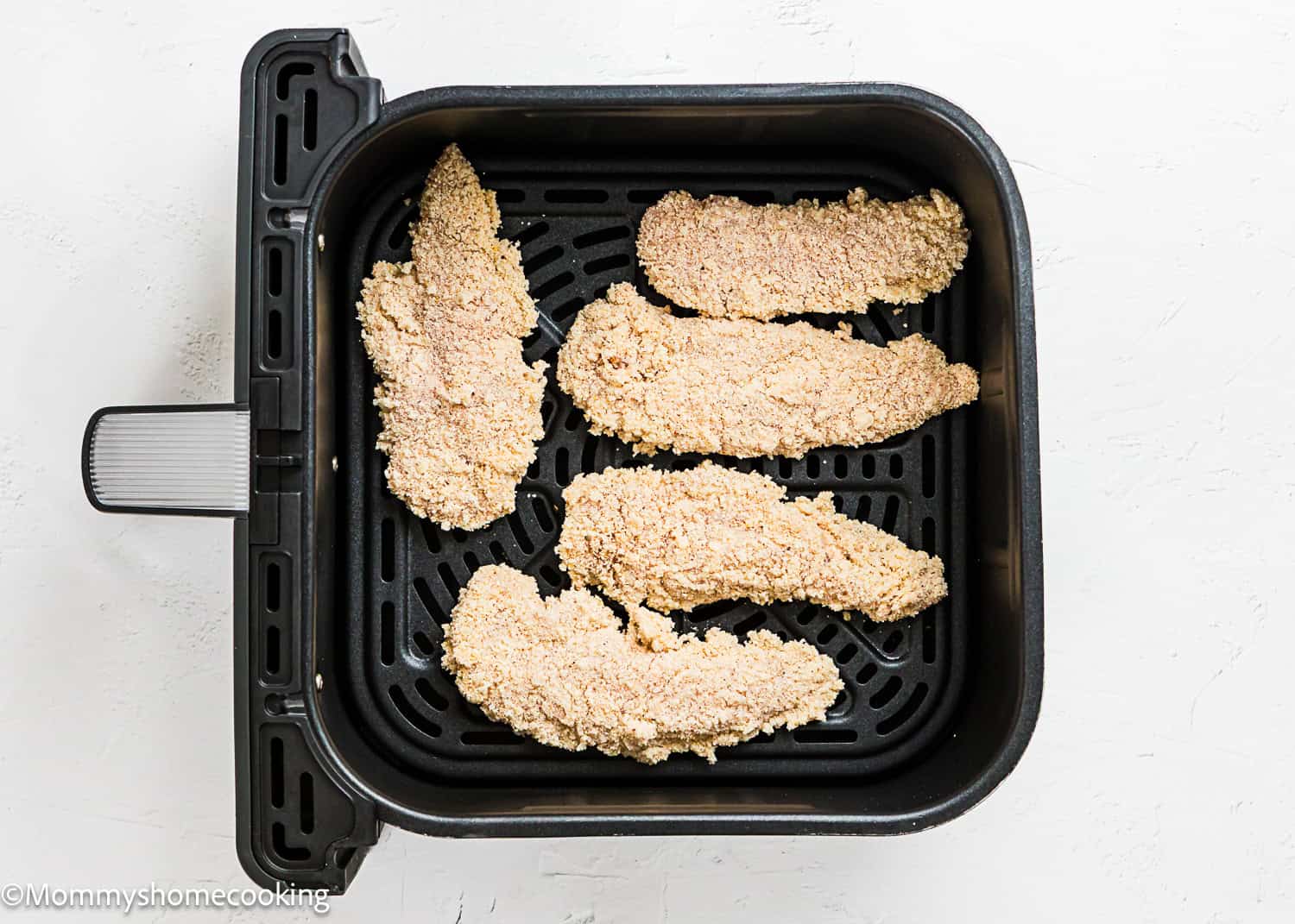 Five breaded chicken tenders, perfect for an Easy Air Fryer Chicken Tender (No Eggs - No Dairy) recipe, are arranged in a single layer inside an air fryer basket on a white surface.