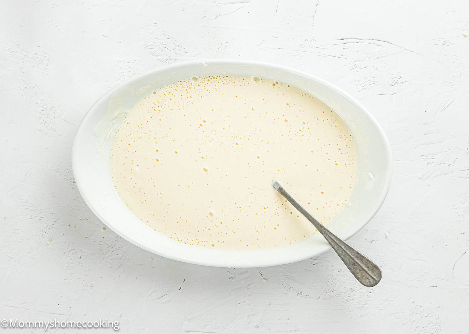 Oval white bowl filled with a creamy, pale yellow batter, perfect for making Easy Air Fryer Chicken Tender (No Eggs - No Dairy), with a metal spoon resting inside, set on a textured white surface.