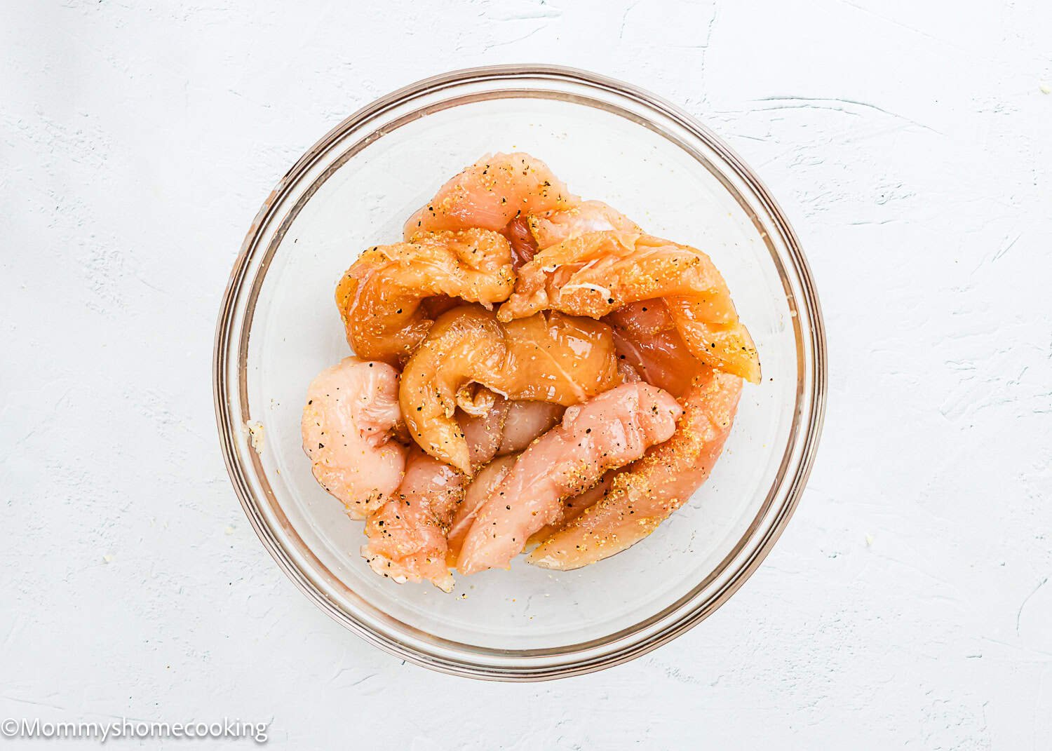 Raw chicken strips seasoned with spices are arranged in a clear glass bowl on a white surface, perfect for making Easy Air Fryer Chicken Tender (No Eggs - No Dairy).