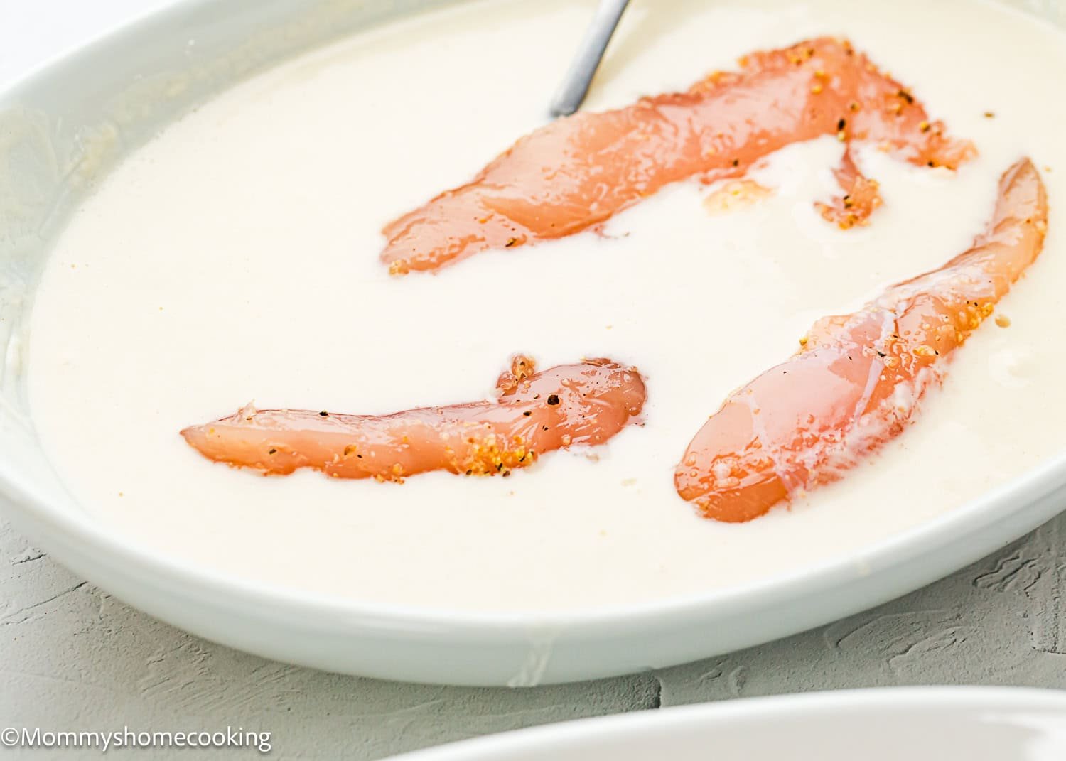 Raw chicken strips marinating in a creamy white sauce inside a shallow white bowl, with a spoon partially visible—perfect for making Easy Air Fryer Chicken Tenders (No Eggs - No Dairy).