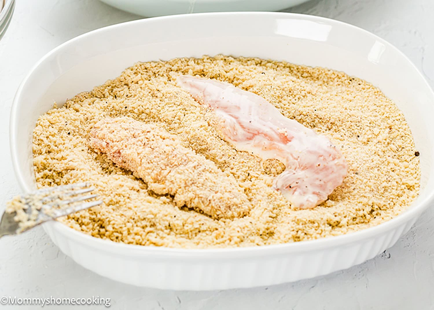 Two pieces of chicken are being coated in breadcrumbs in a white dish—one piece is partially covered, and the other is fully coated—perfect for making Easy Air Fryer Chicken Tenders with no eggs and no dairy.