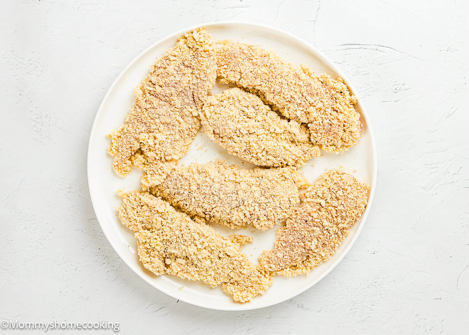 A white plate holds six uncooked, breaded Easy Air Fryer Chicken Tenders (No Eggs - No Dairy) on a light textured surface.
