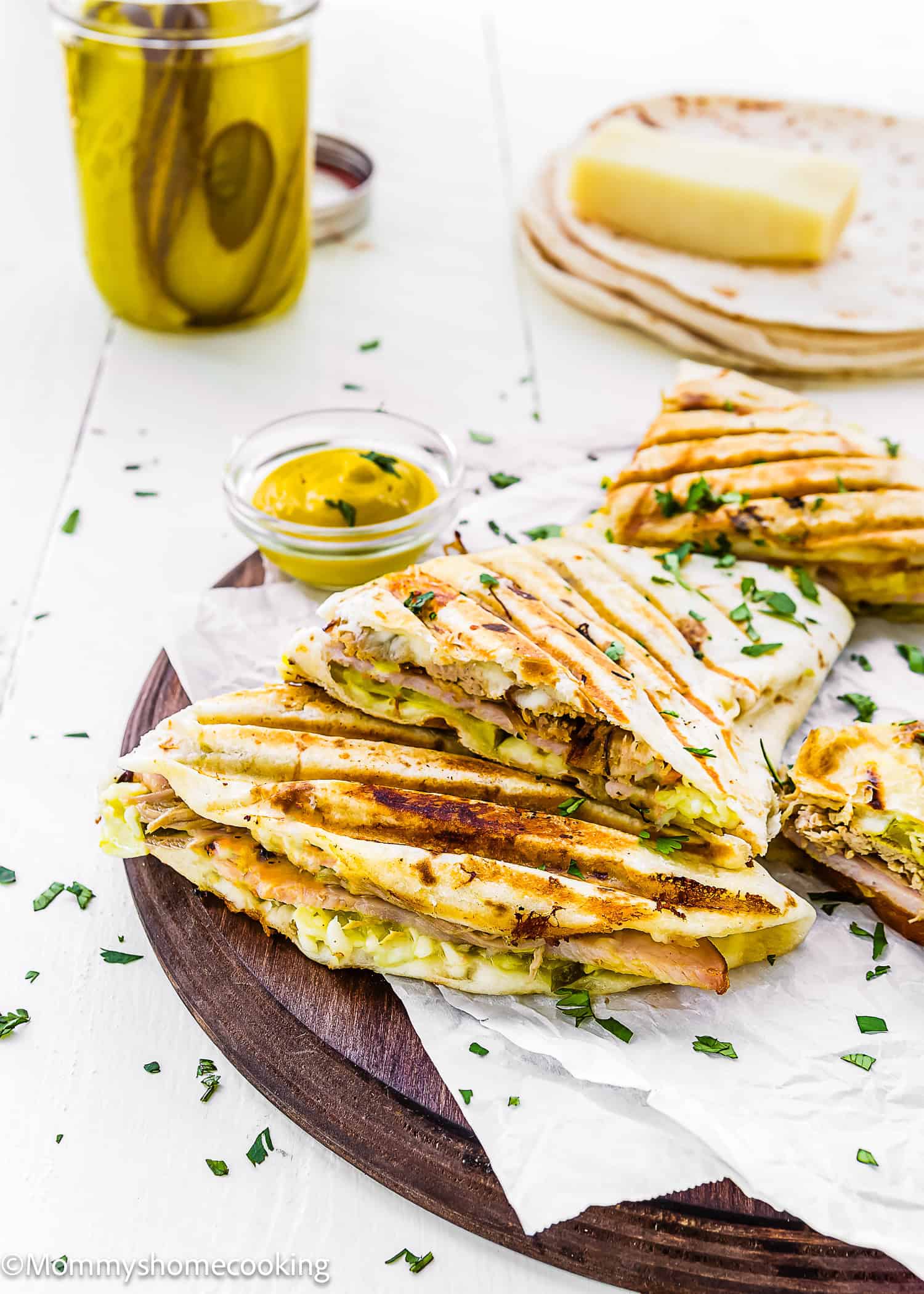 Grilled chicken paninis with melted cheese and herbs are stacked on a wooden board, inspired by Easy Cuban Quesadillas, with mustard, sandwich bread, and a cheese block in the background.